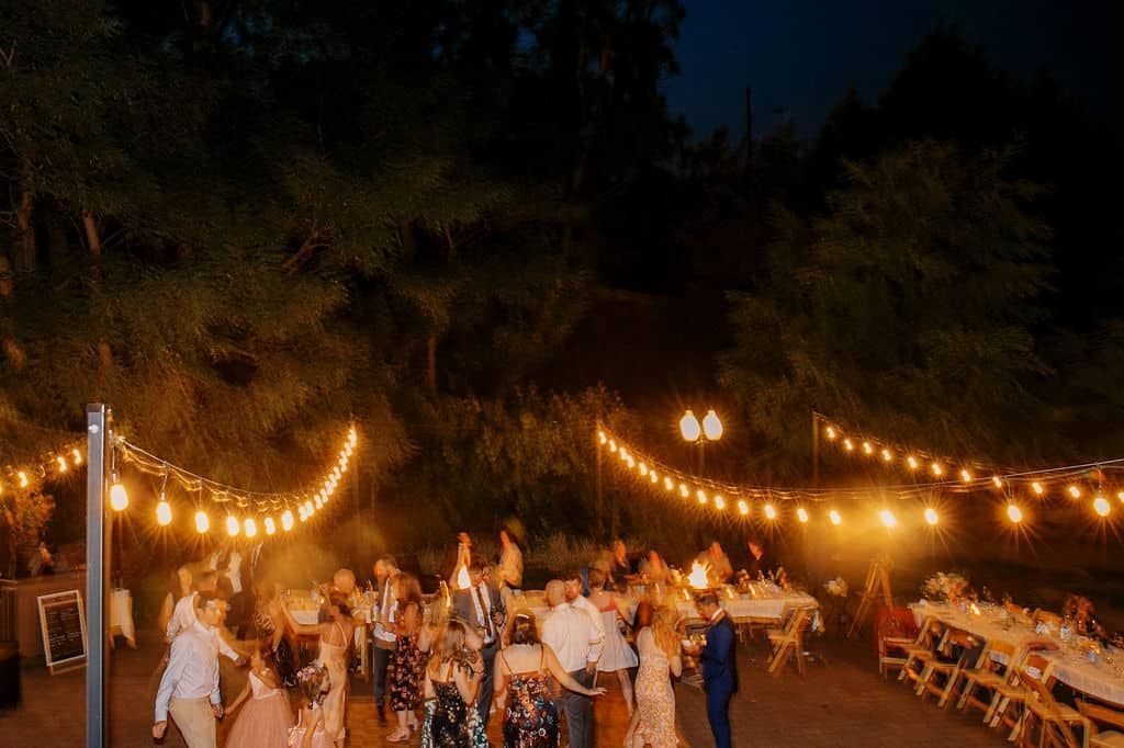 A group of people are dancing at a wedding reception under a string of lights.