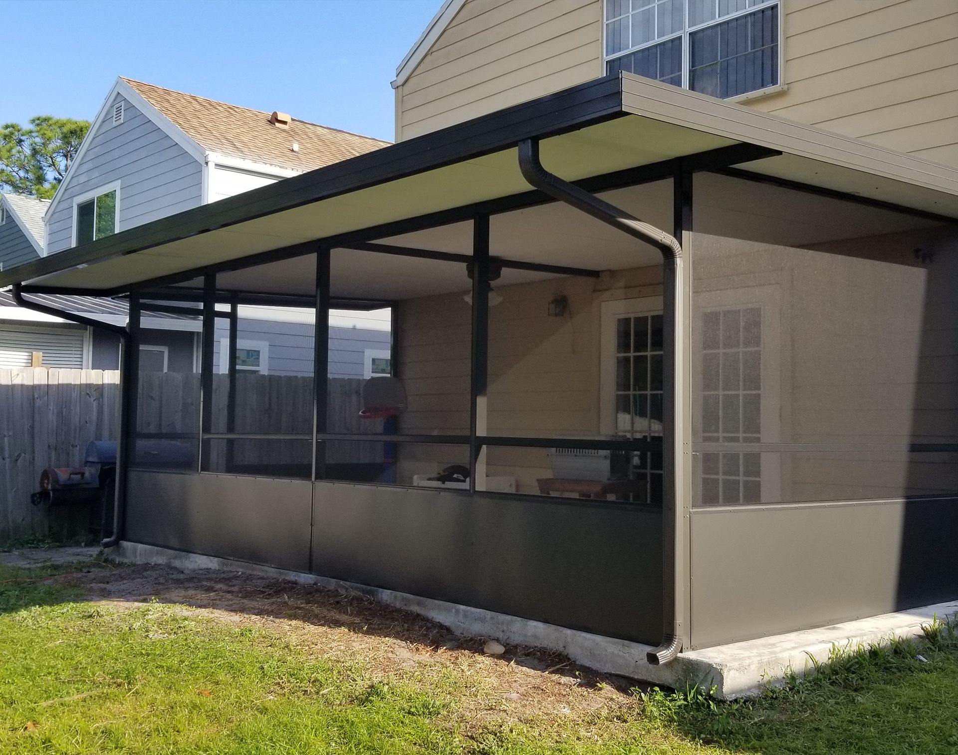 Screened-in porch attached to a beige house, with dark trim and roof, and a green lawn in front.