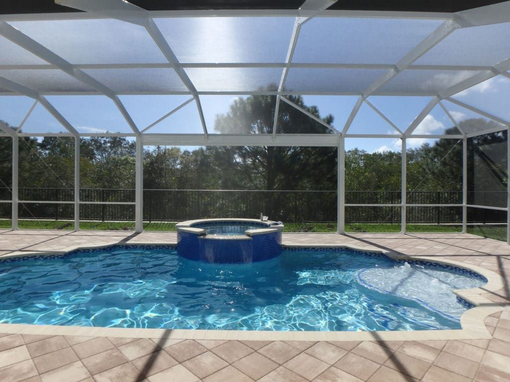 Pool enclosed in a white screen with a hot tub and blue water. Lush green trees in the background.