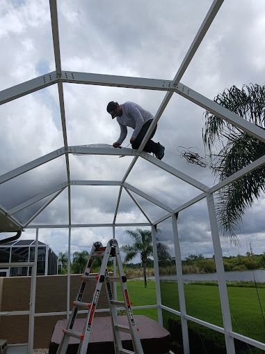 Man on a white screen enclosure roof, working. Ladder below. Cloudy sky, green lawn, and water in the background.