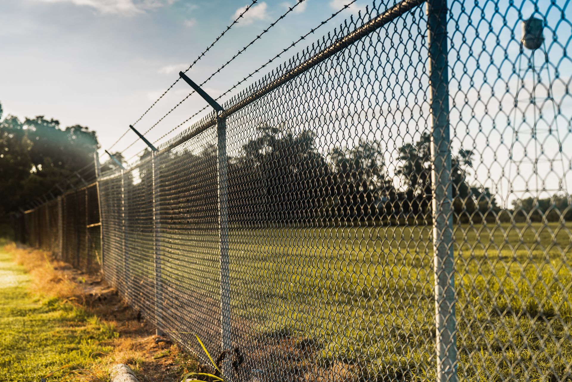 A tall chain-link fence topped with barbed wire runs diagonally through a sunny, grassy field.