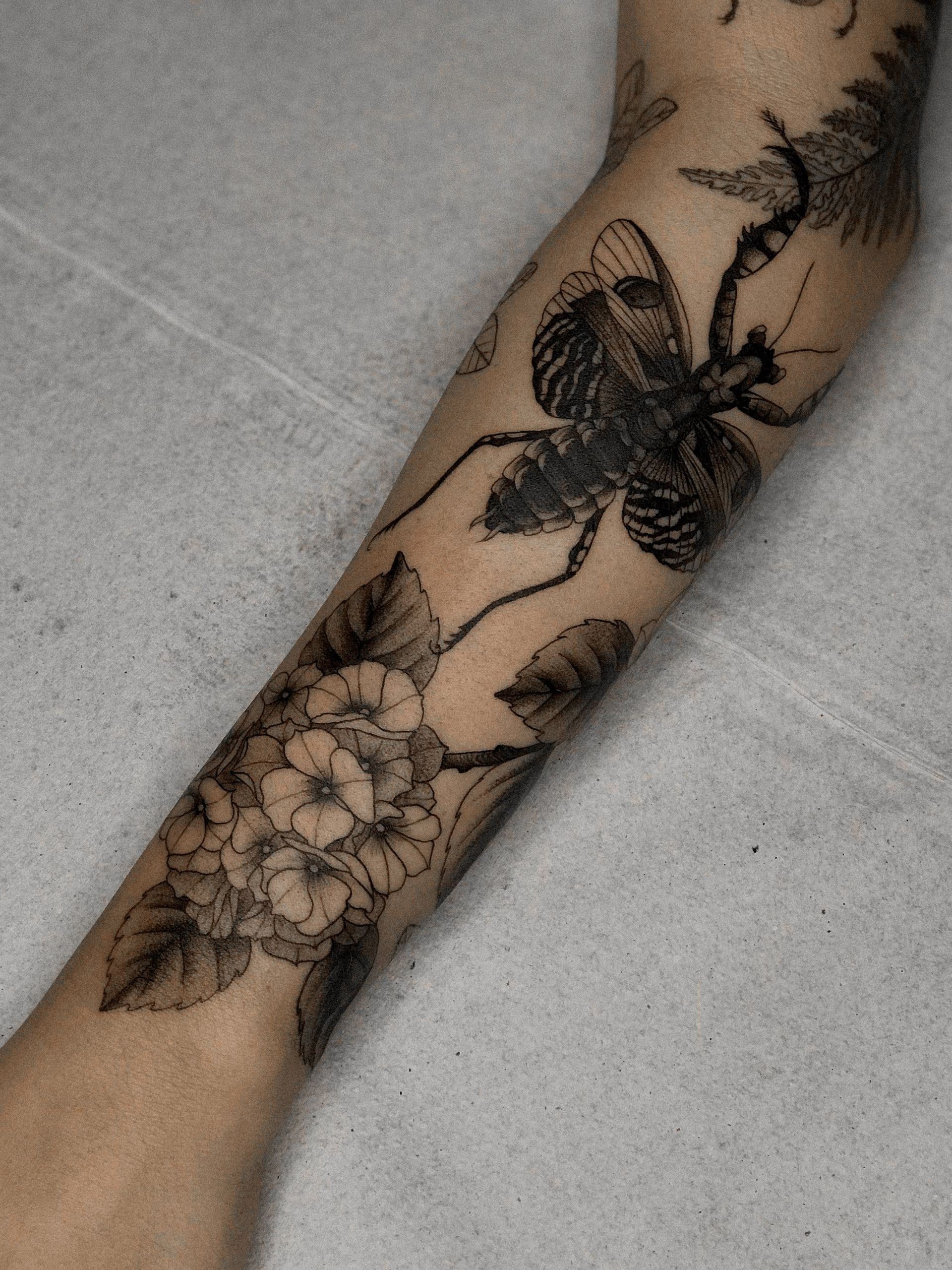 Uma tatuagem de um louva-a-deus e flores no braço de uma pessoa
