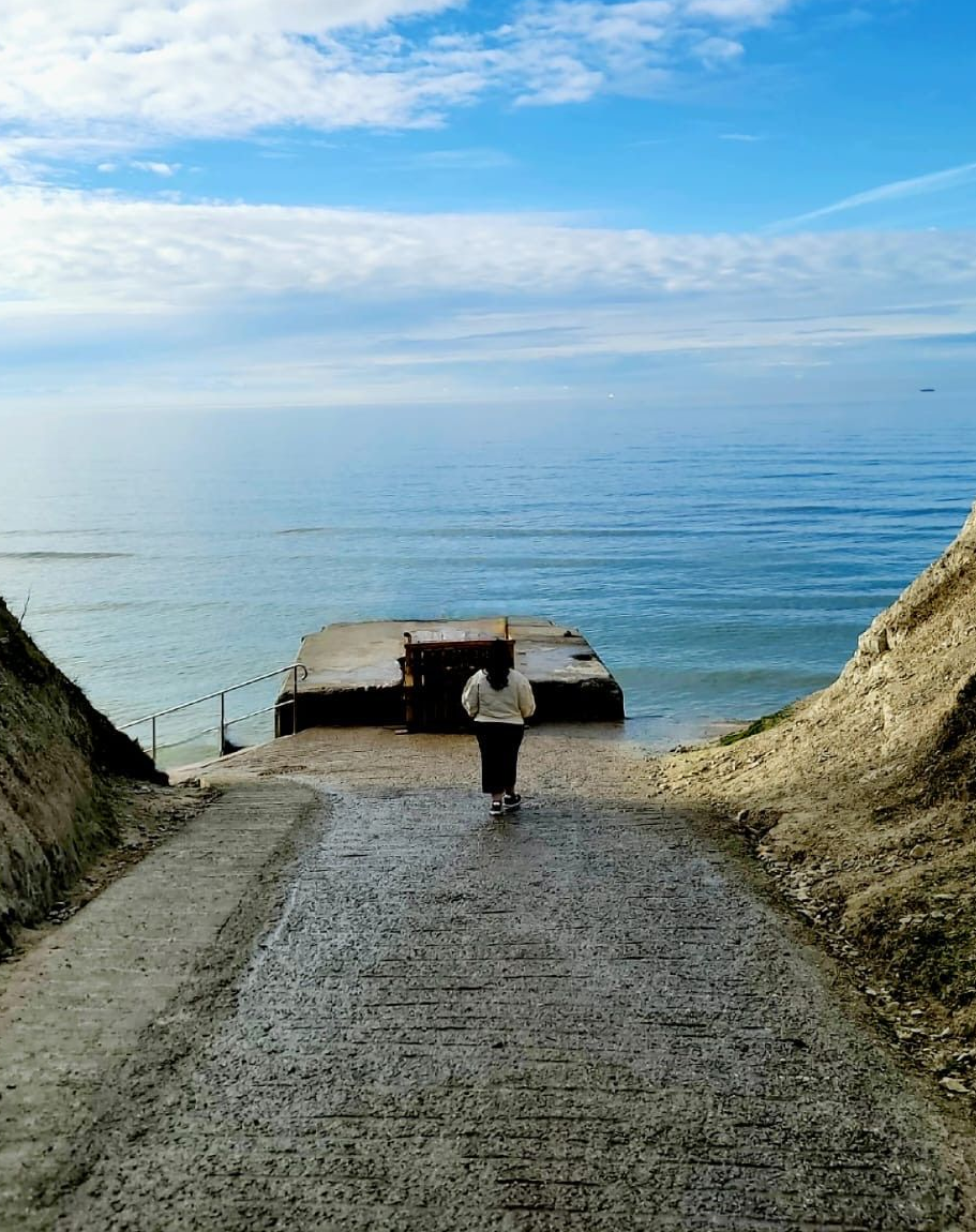 Person stands at a concrete ramp leading to a weathered platform on the ocean. Blue sky, water, and cliffs.