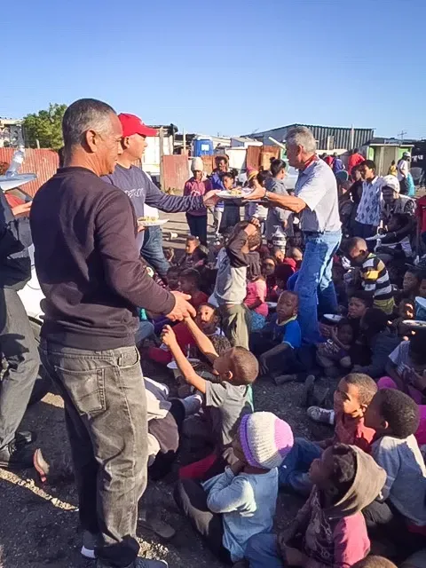 Men serving food to a group of children, seated outside in a community setting.