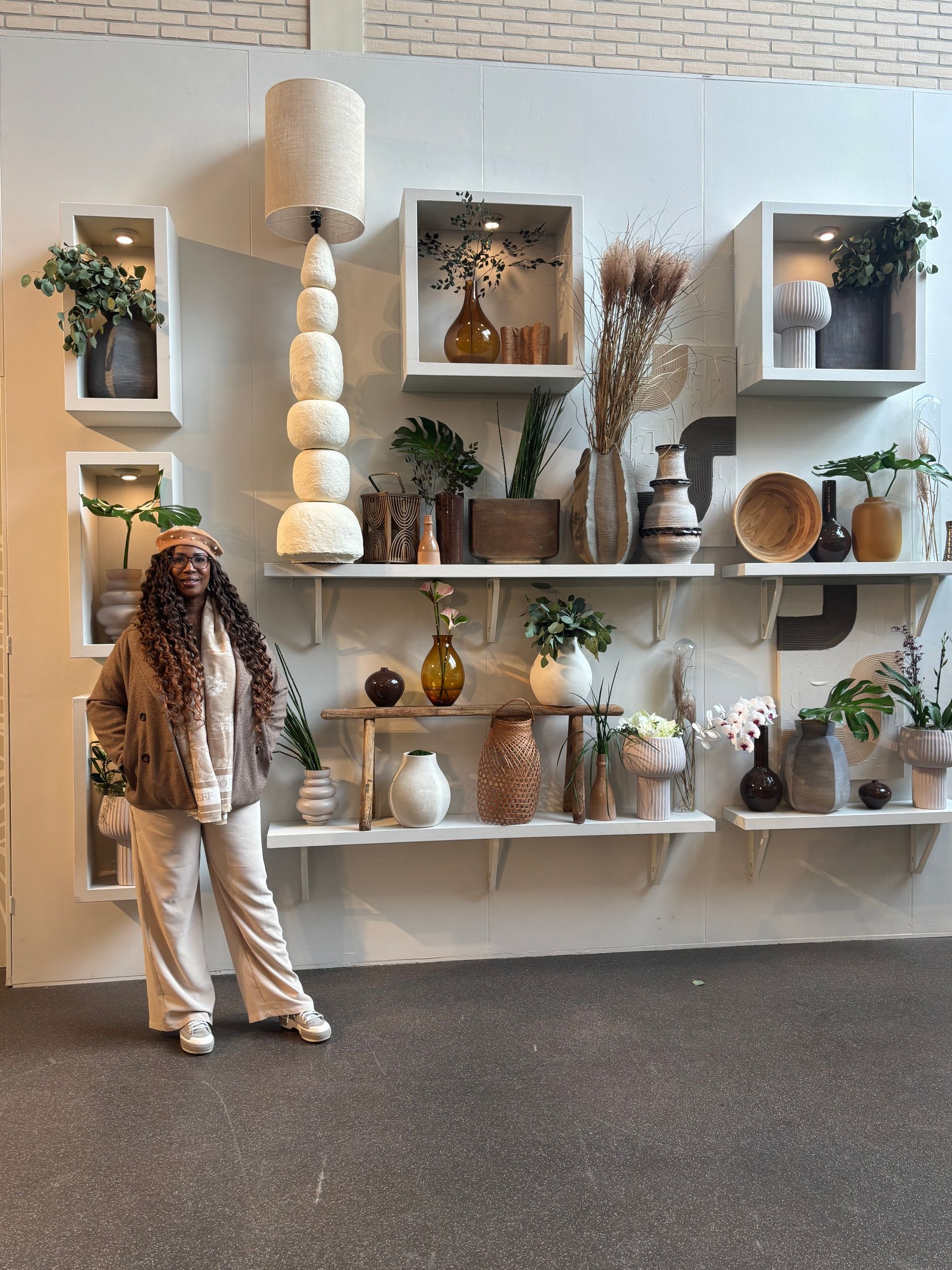 Woman stands by a display of shelves with vases and plants, a tall lamp, and square wall decor.