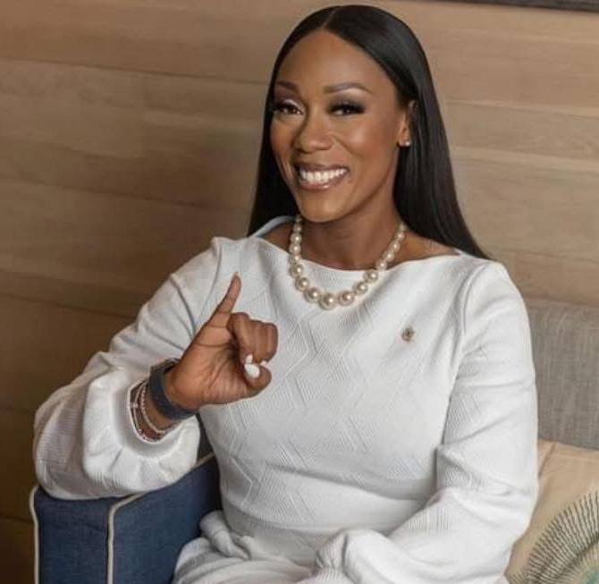 Woman in white dress, pearl necklace, smiling, holding up finger, in a well-lit setting.