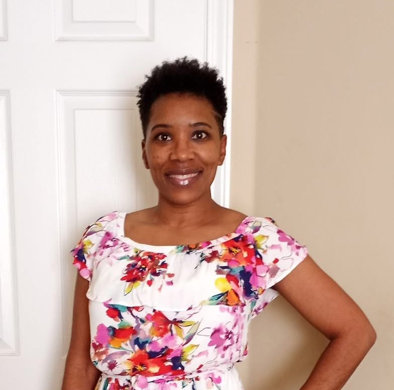 Woman with short dark hair, wearing floral dress, smiling in front of a white door.
