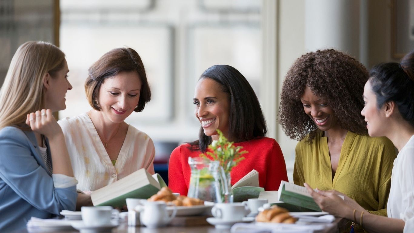 Five women at a table, smiling and reading books. Light and airy setting, food and drinks present.