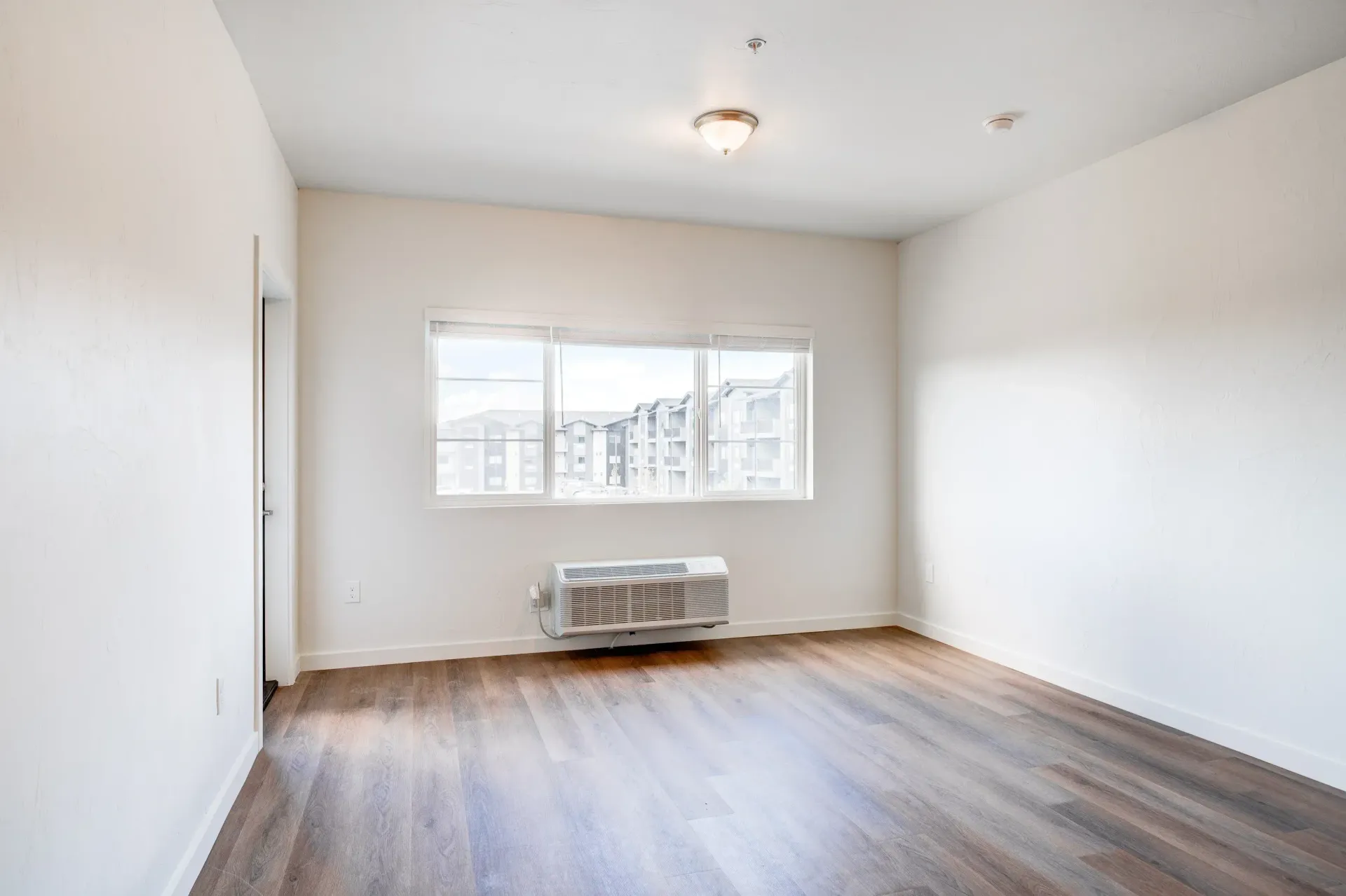 Empty apartment room with window and wall-mounted air conditioner; wood-like flooring.