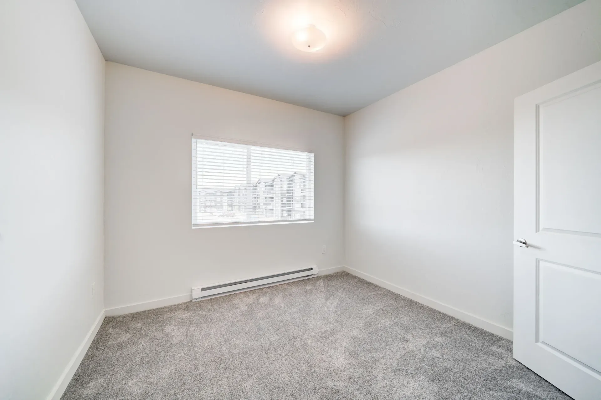 Empty bedroom with white walls, a window with blinds, carpet, and a closed white door.