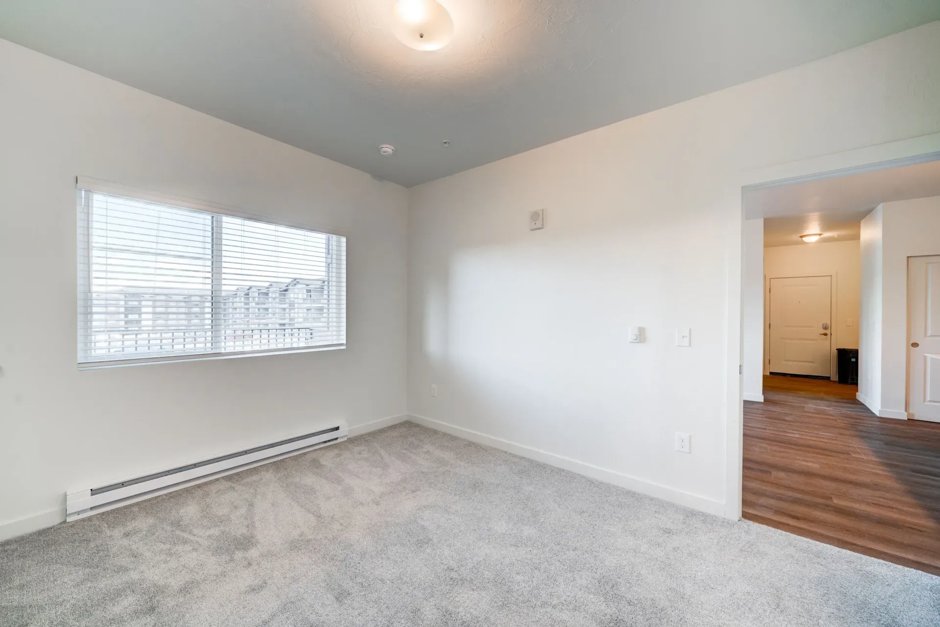 Empty living room with a large window, white walls, light carpet, baseboard heater, and doorway to a hallway.