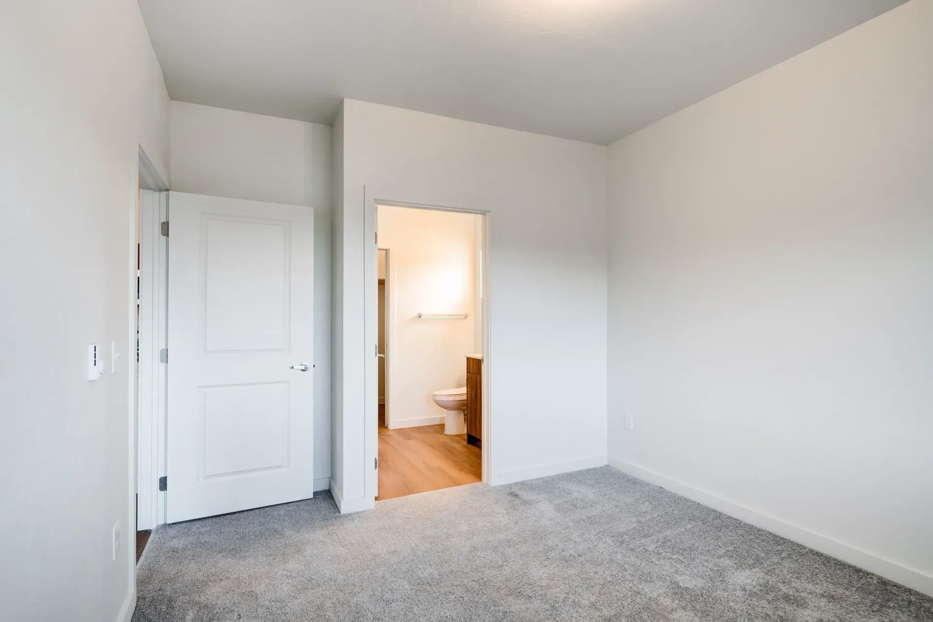 Empty bedroom with white walls, gray carpet, and an open doorway to a bathroom.