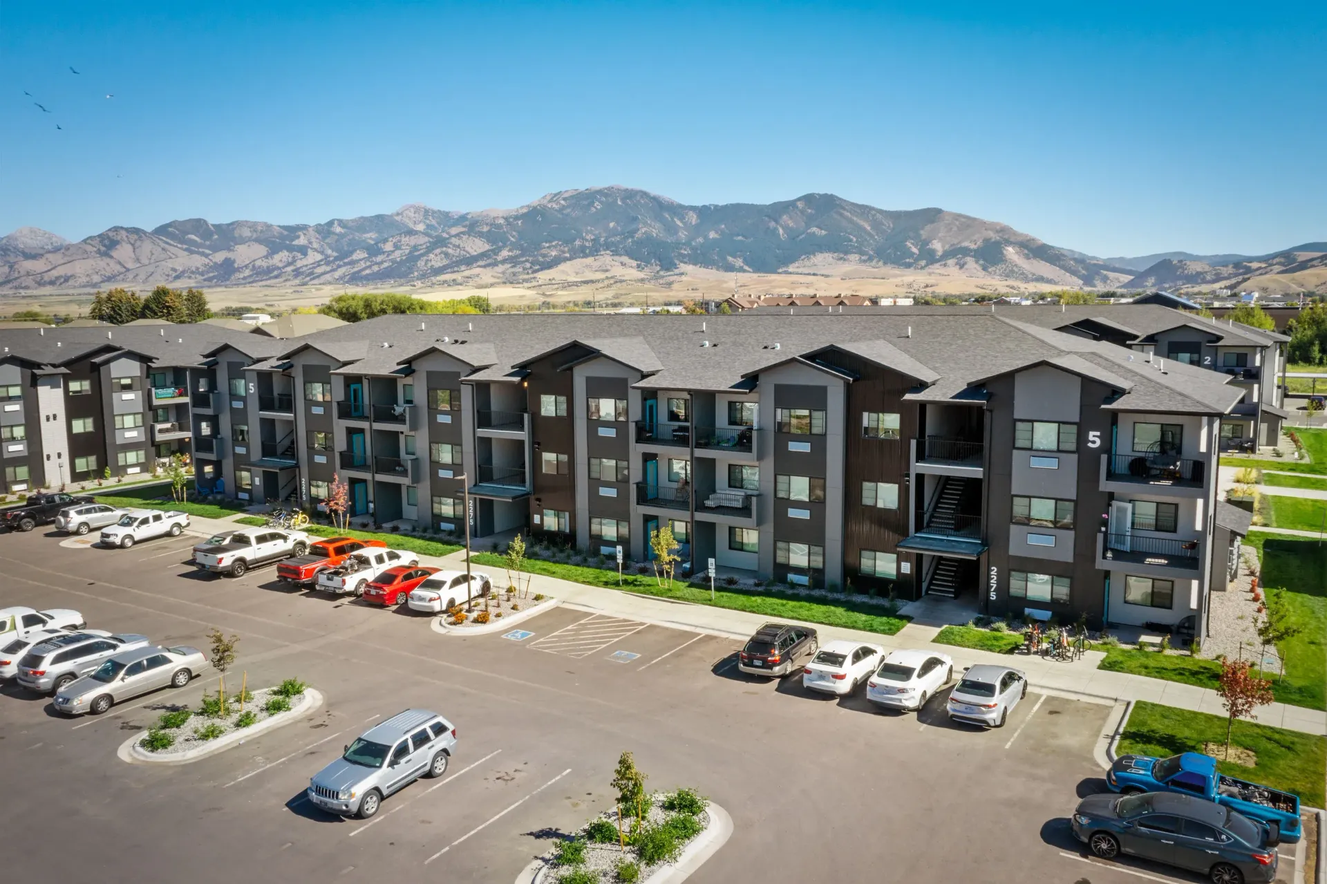 Aerial view of a modern apartment complex with multiple buildings, parking, and distant mountains.