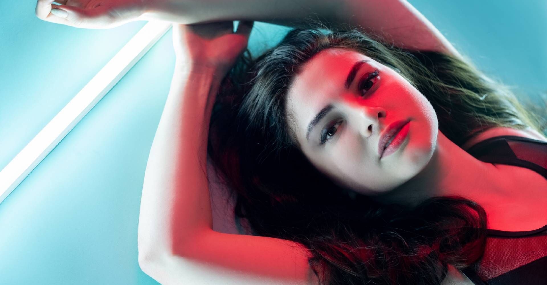 Woman with dark hair reclines under red and blue lights, arms raised, looking up.