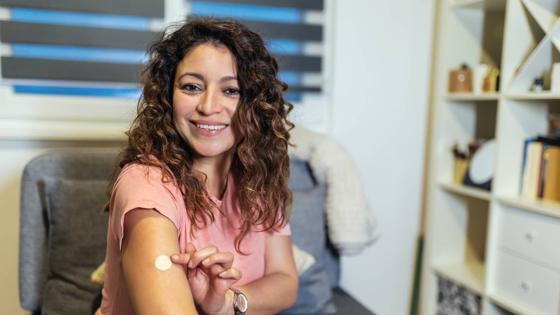 Woman applying cream to her arm, smiling. Indoors, next to a chair and shelf.