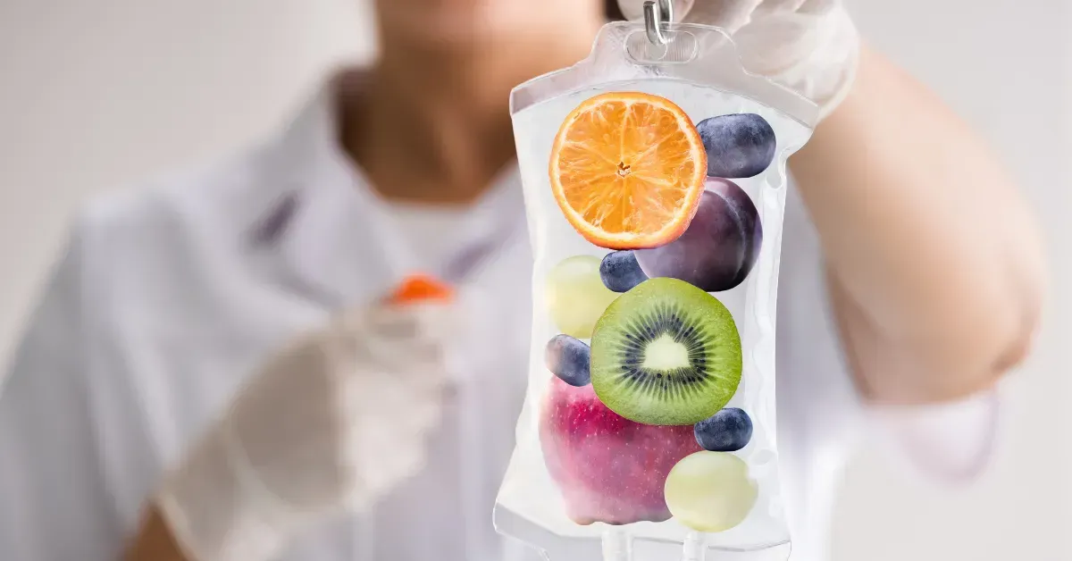 Person in white lab coat holding IV bag filled with fruit slices: orange, kiwi, apple, berries.