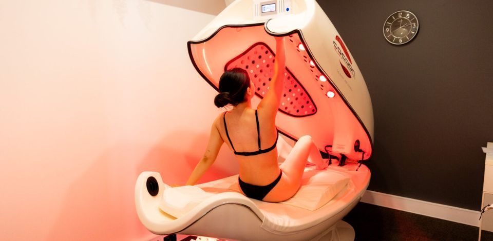 Woman in swimsuit inside a red light pod, raising arm. White and gray room, clock on wall.