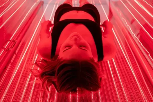 Woman in black activewear inside a red light therapy device, looking down with eyes closed.