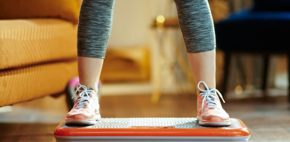 Person exercising on a vibrating fitness platform, wearing sneakers and grey leggings, indoors.