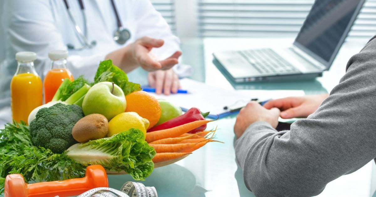 Doctor consulting a patient about nutrition, with fresh produce, juice, weights, and a laptop on the desk.