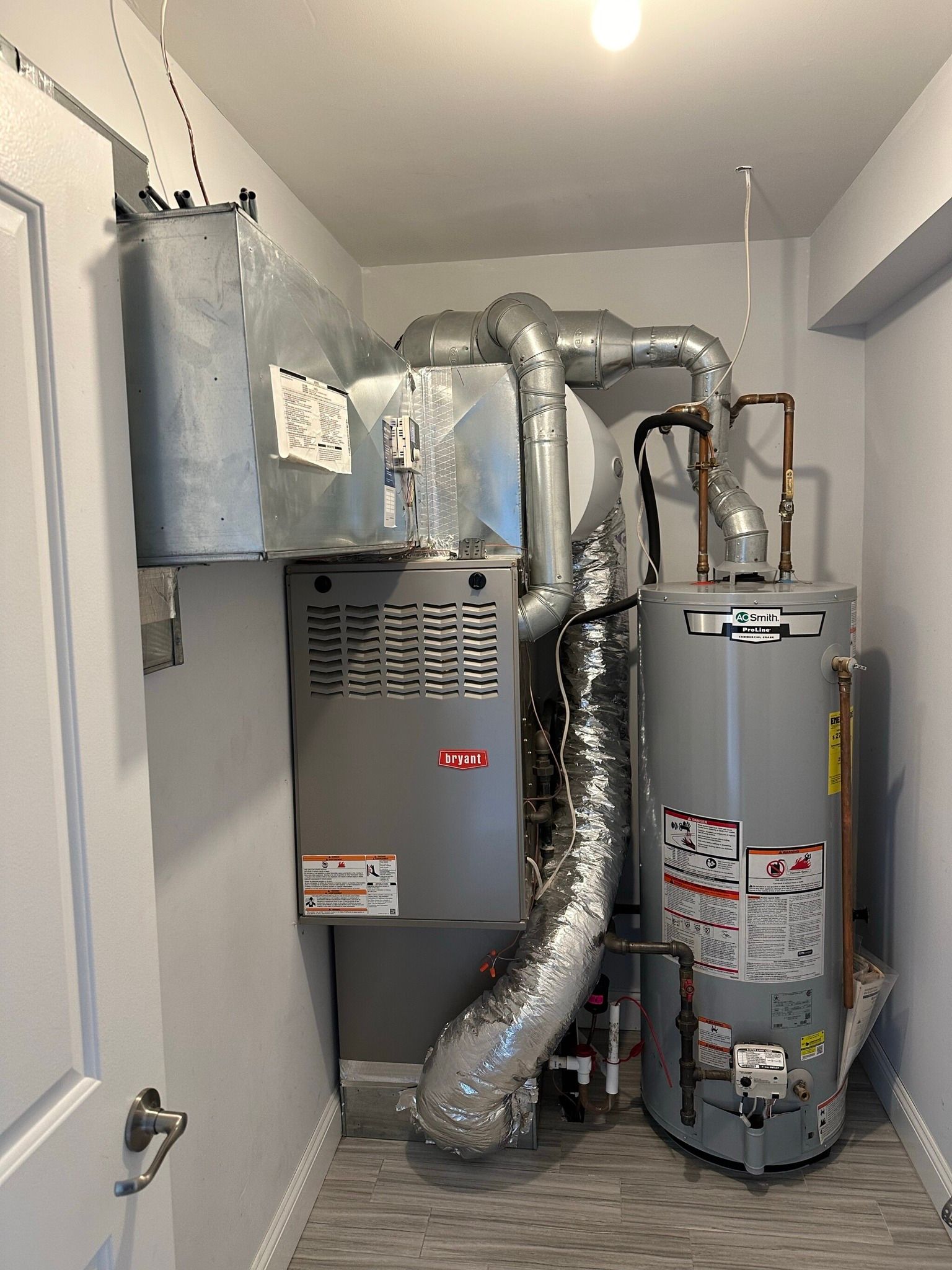 Furnace and water heater in a small utility closet. Gray metal appliances, silver ducts, and white walls.