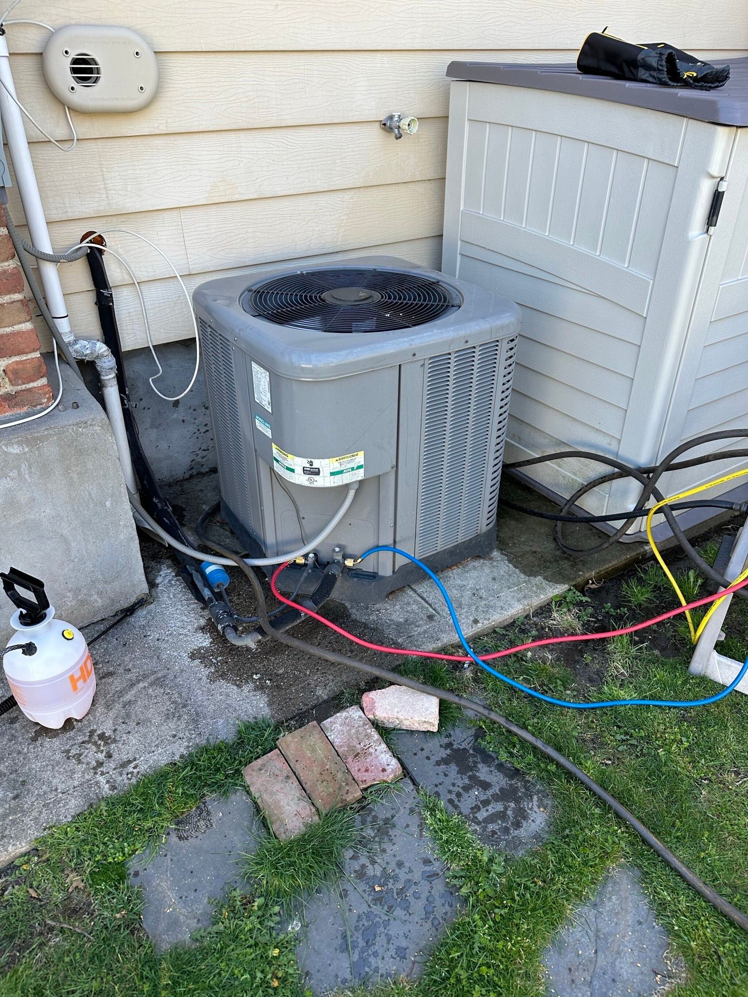 An air conditioning unit being serviced outdoors; hoses attached, gray and blue.