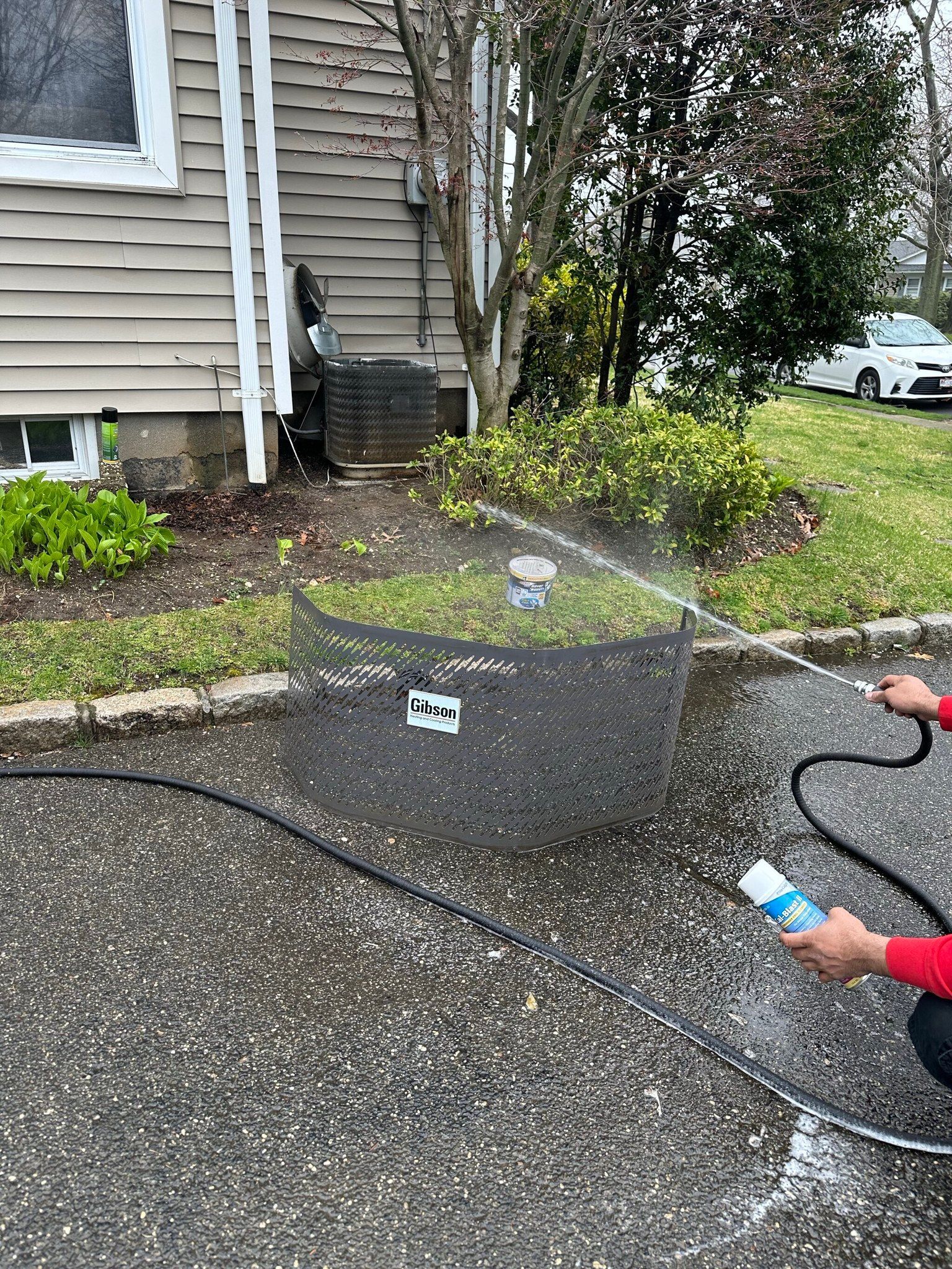Person sprays a dark circular container with water near a house and bushes.