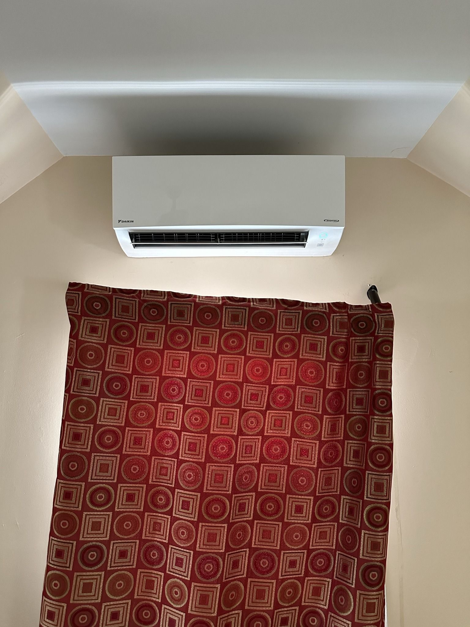 White air conditioner mounted on a beige wall above a patterned red curtain.