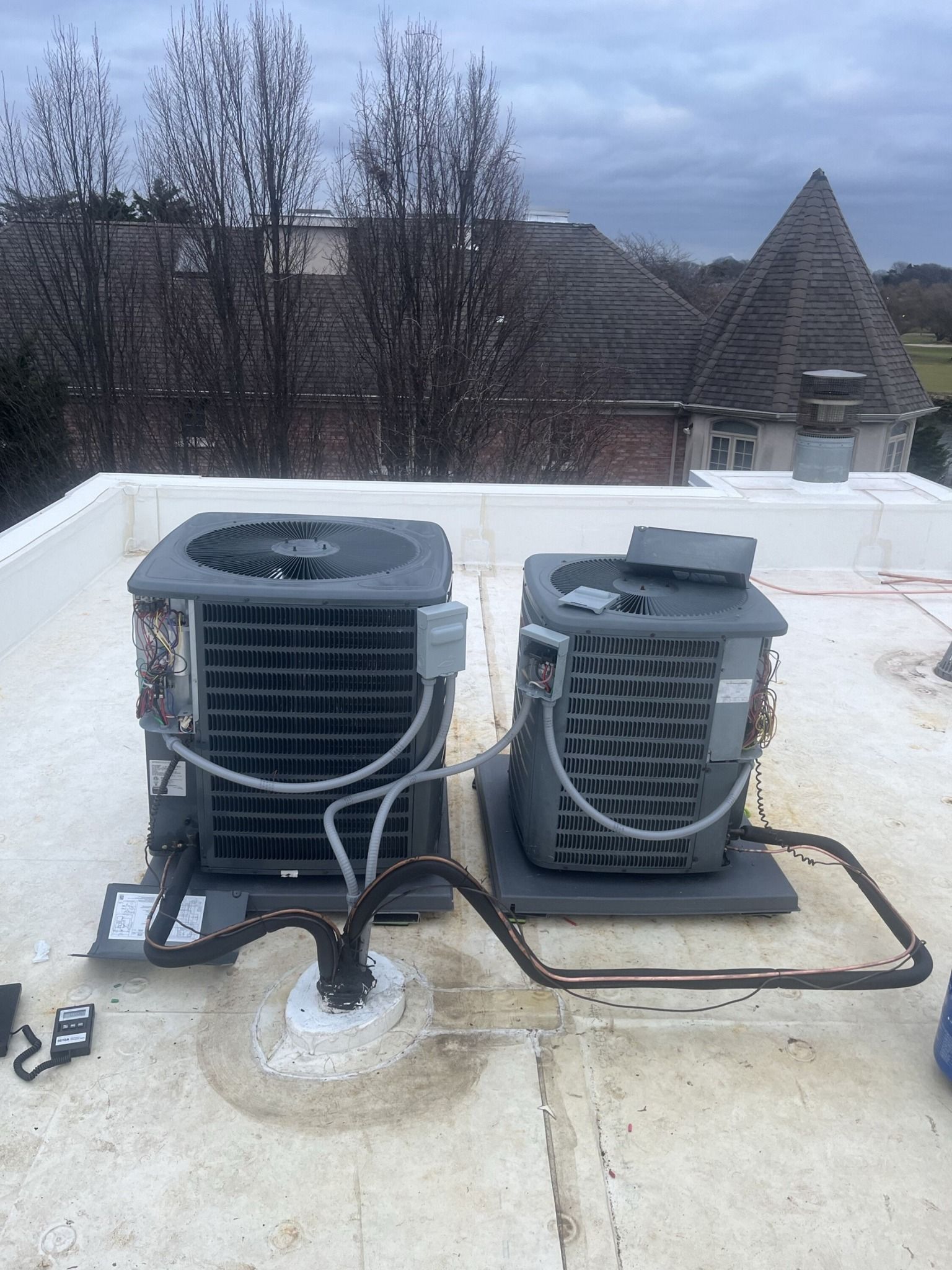 Two gray air conditioning units on a white rooftop, connected by black pipes.