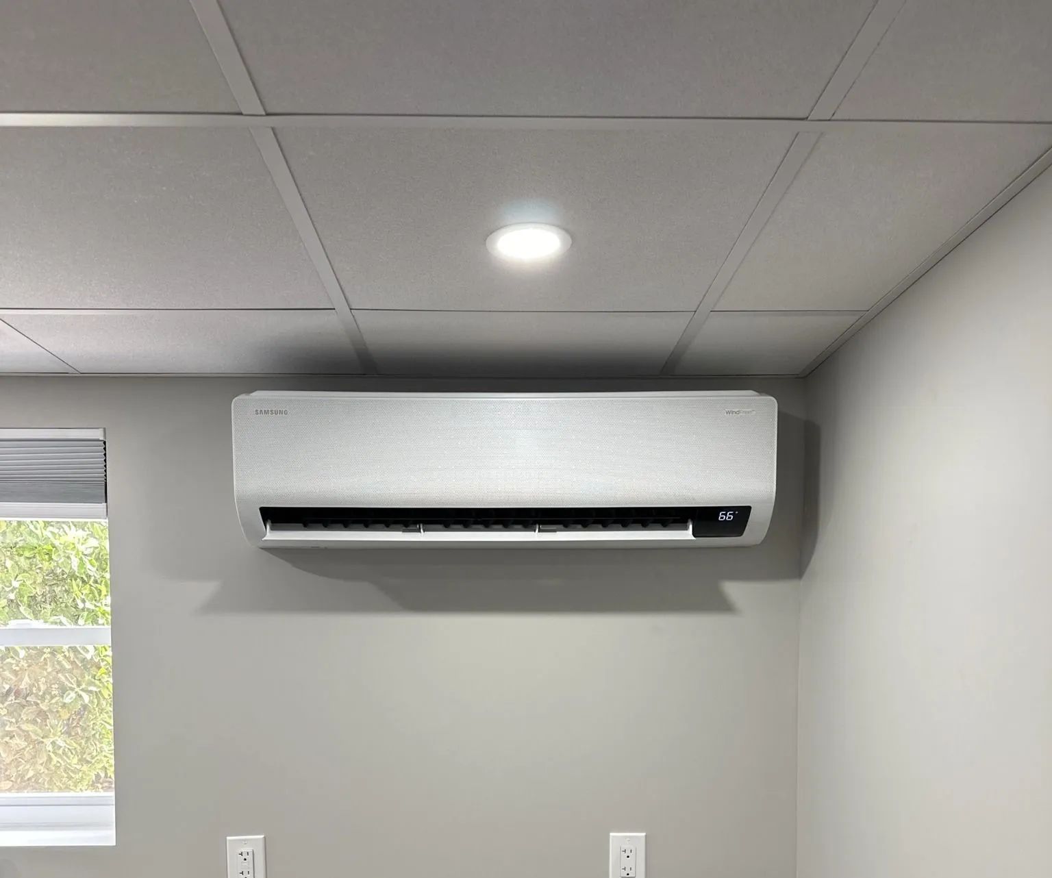 White wall-mounted air conditioner on a gray wall. A ceiling light and window are also visible.