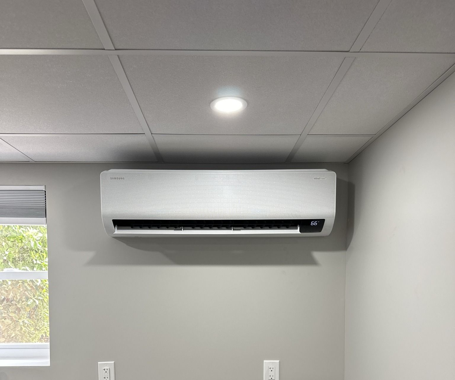 White wall-mounted air conditioner on gray wall below a ceiling light.