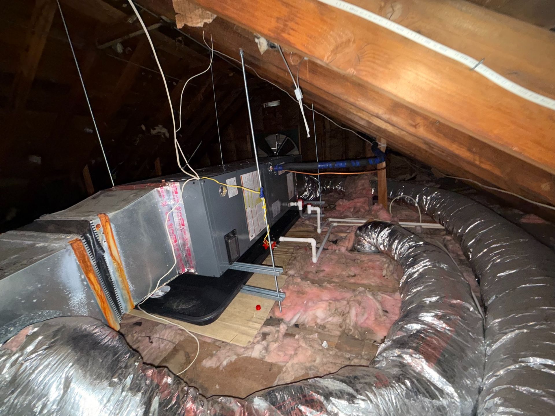 HVAC unit in attic, surrounded by ductwork and insulation. Exposed wooden beams overhead.