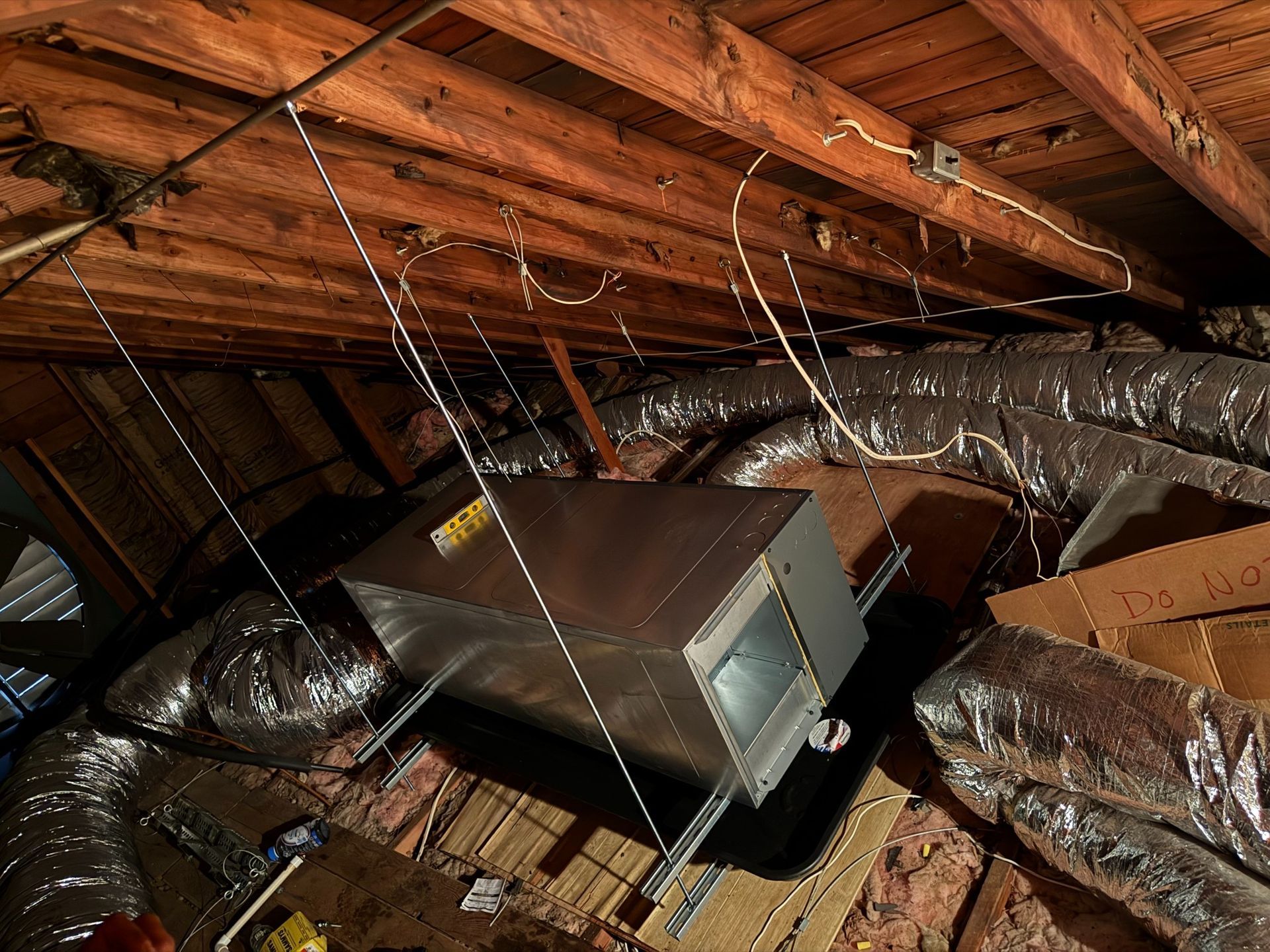 HVAC unit in a dark attic, suspended from wooden beams by wires, surrounded by ducts and insulation.