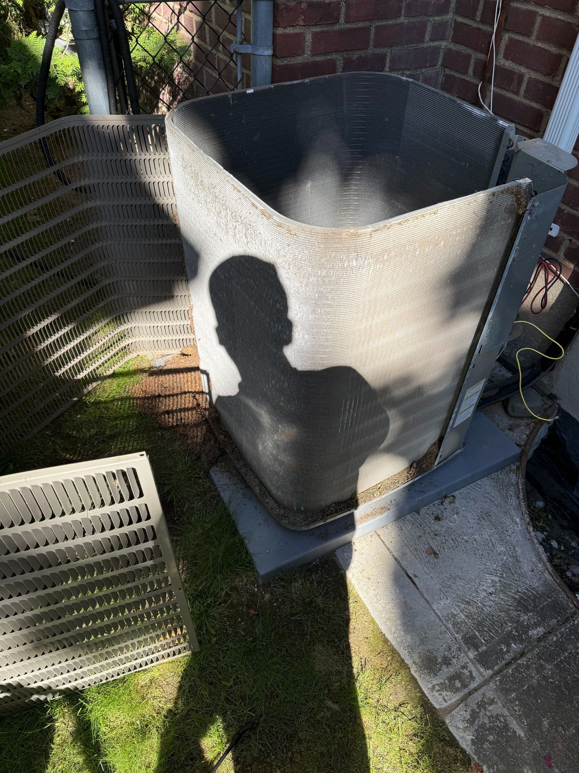 A person's shadow cast on a central air conditioning unit in a yard; one grille panel is detached.
