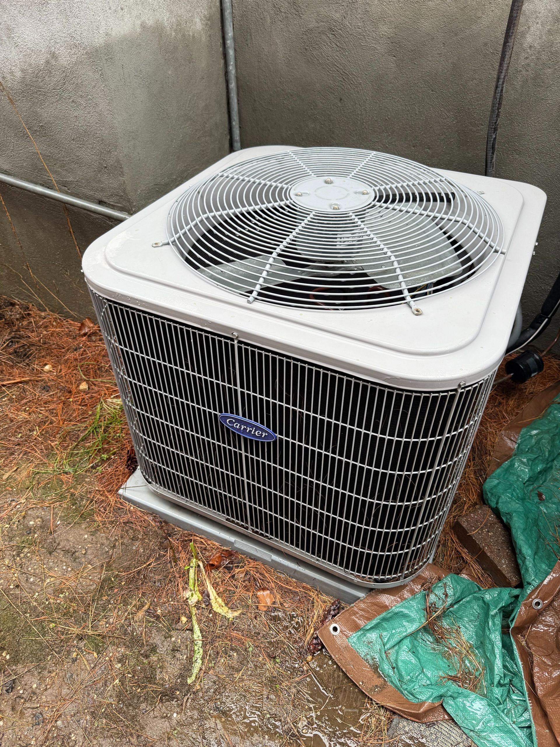 Air conditioning unit outside, gray metal, fan on top, black metal sides, gray concrete wall in background.