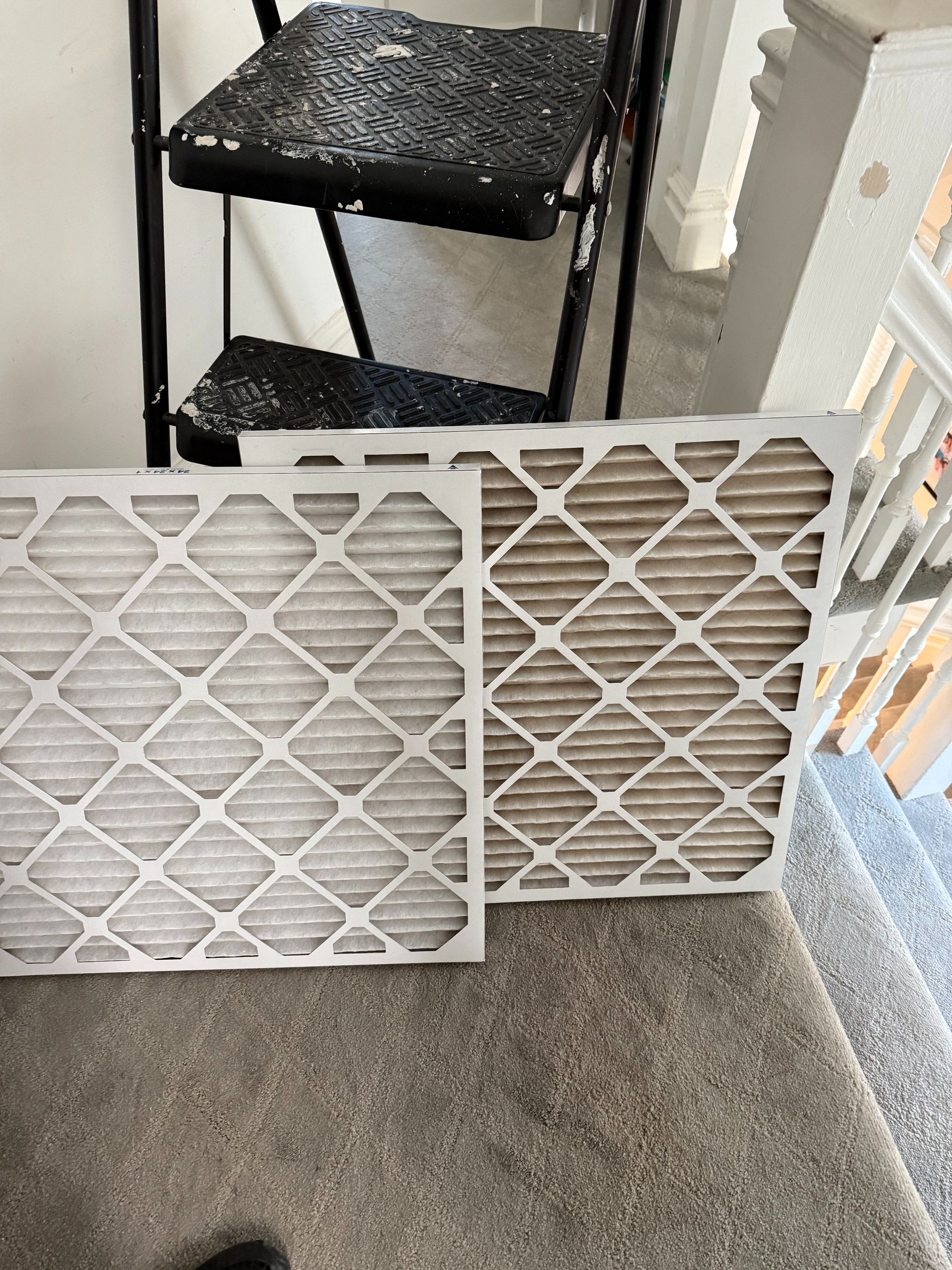 Two air filters: clean white one next to a dirty brown one, on carpeted steps with a stepladder.