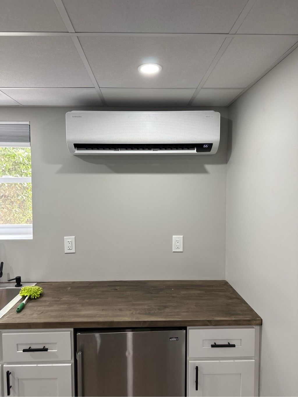 Air conditioning unit mounted above a countertop with cabinets and a mini fridge. Gray walls, white ceiling.