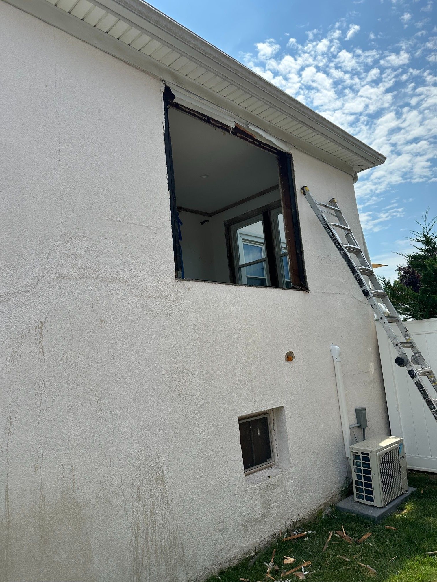 Exterior house wall with a large window frame, a smaller window, and a ladder, against a blue sky.