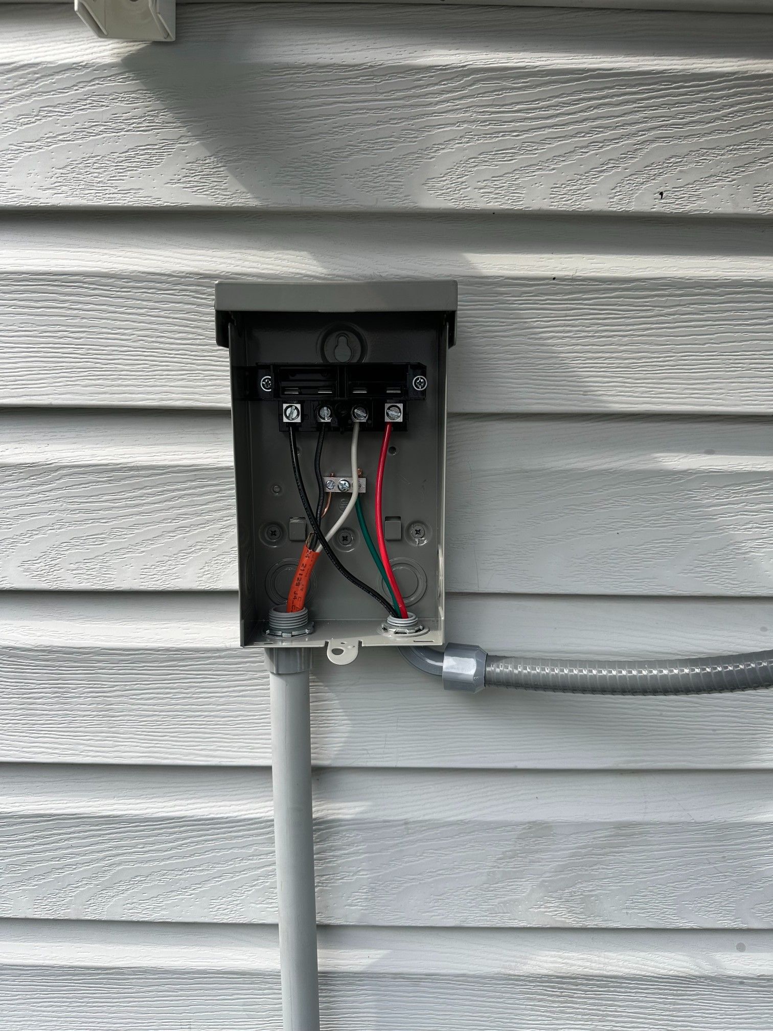 Metal electrical box mounted on gray siding, wires connected inside. Gray conduit runs down and to the side.