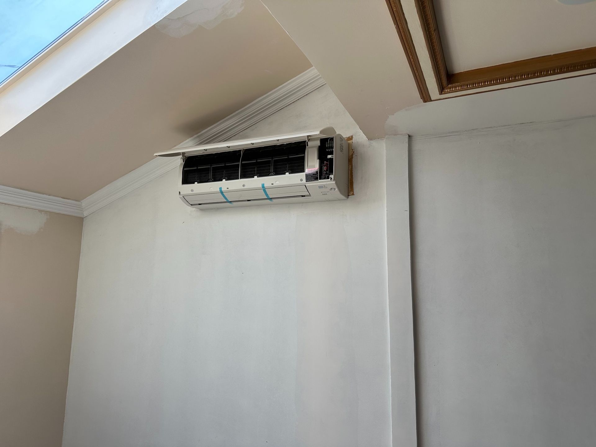 White air conditioner mounted on a white wall, under a white and cream ceiling.