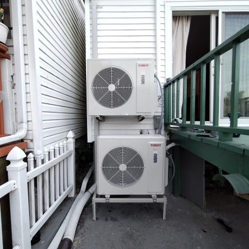 Two white air conditioning units stacked on a metal stand next to a deck and house.