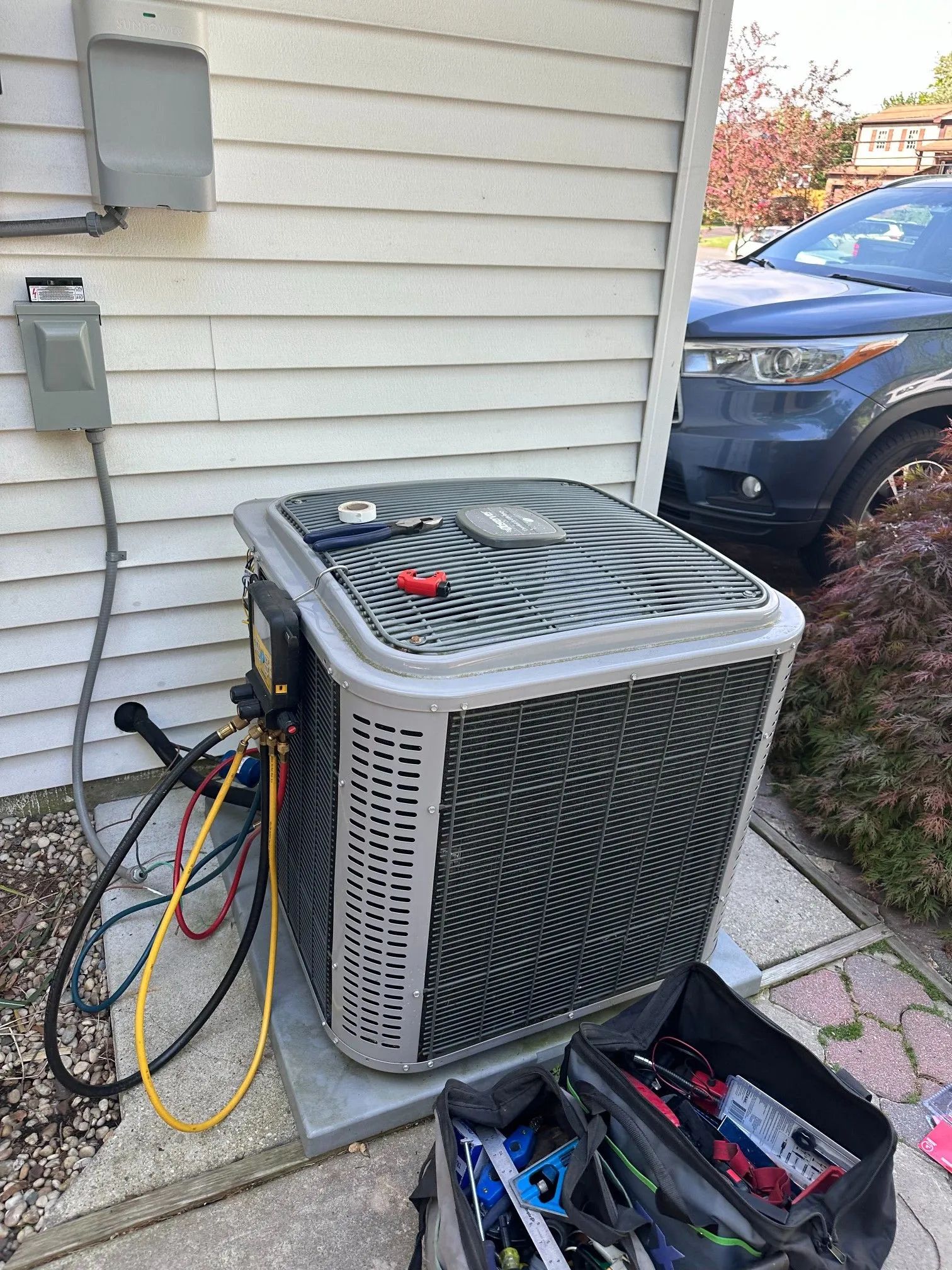 HVAC unit with hoses connected, next to a house with a car parked nearby and a toolbox in the foreground.