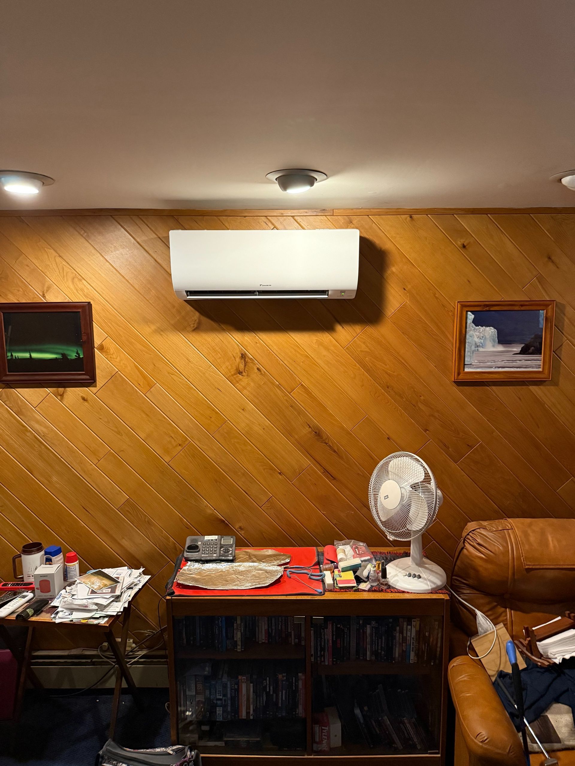 Air conditioner mounted on wood-paneled wall, flanked by framed art, above a console with items and a fan.