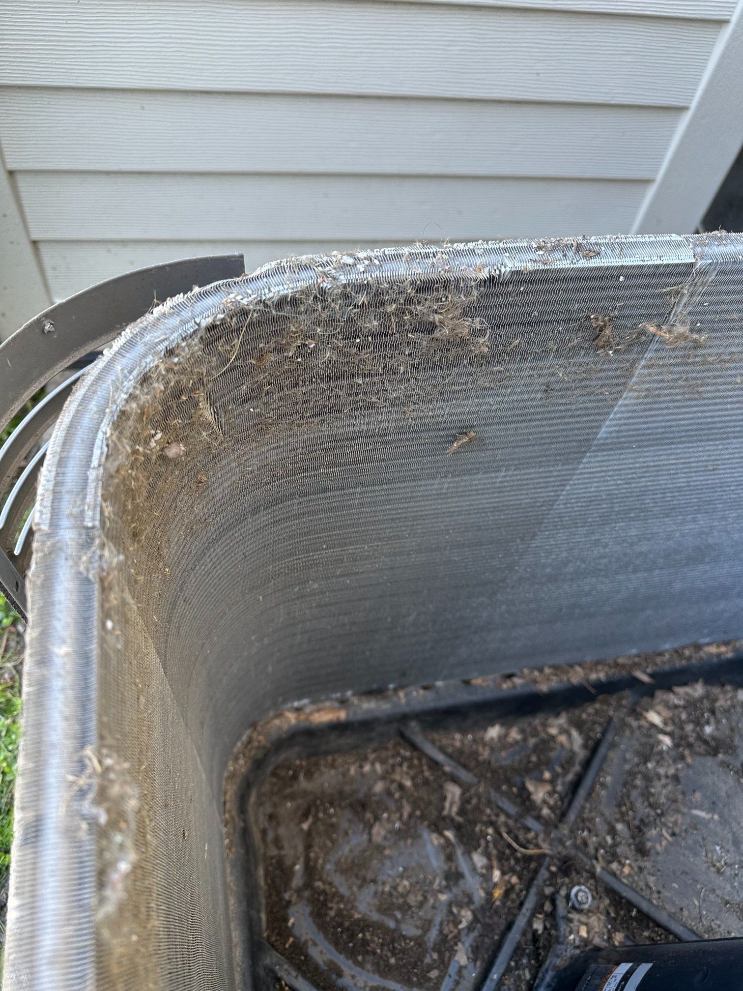 Dirty air conditioning unit, close-up view. Coils covered in dirt and debris. Outdoor setting with siding visible.