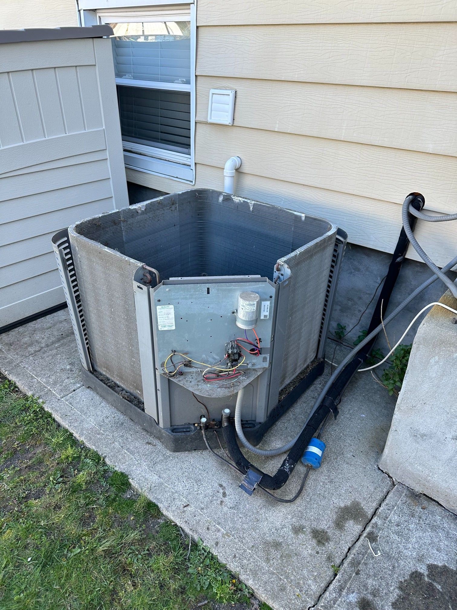 Exterior view of a damaged hexagonal air conditioner. Exposed internal components, near a beige wall and concrete.