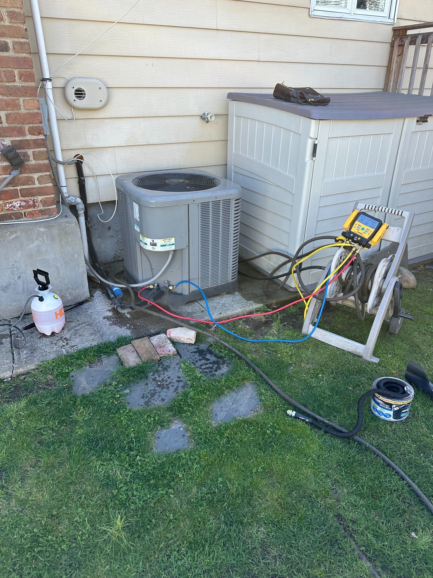 AC unit being serviced outdoors with gauges, hoses, and a spray bottle on grass.