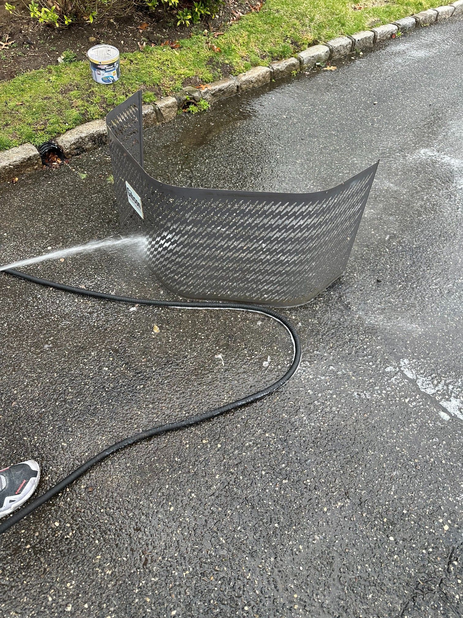 A person washes a black well window cover on a wet driveway with a pressure washer.