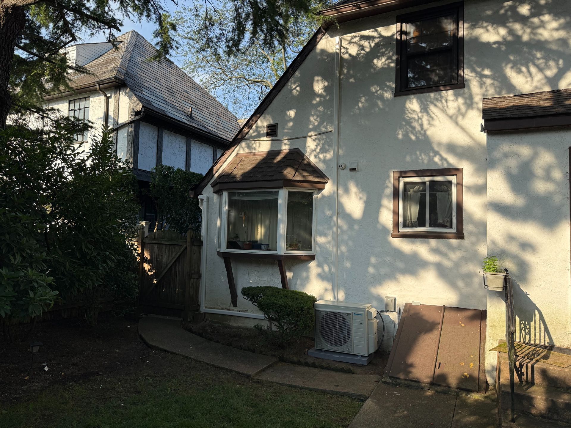Side view of a cream-colored house with a bay window and a smaller window, nestled near trees on a sunny day.