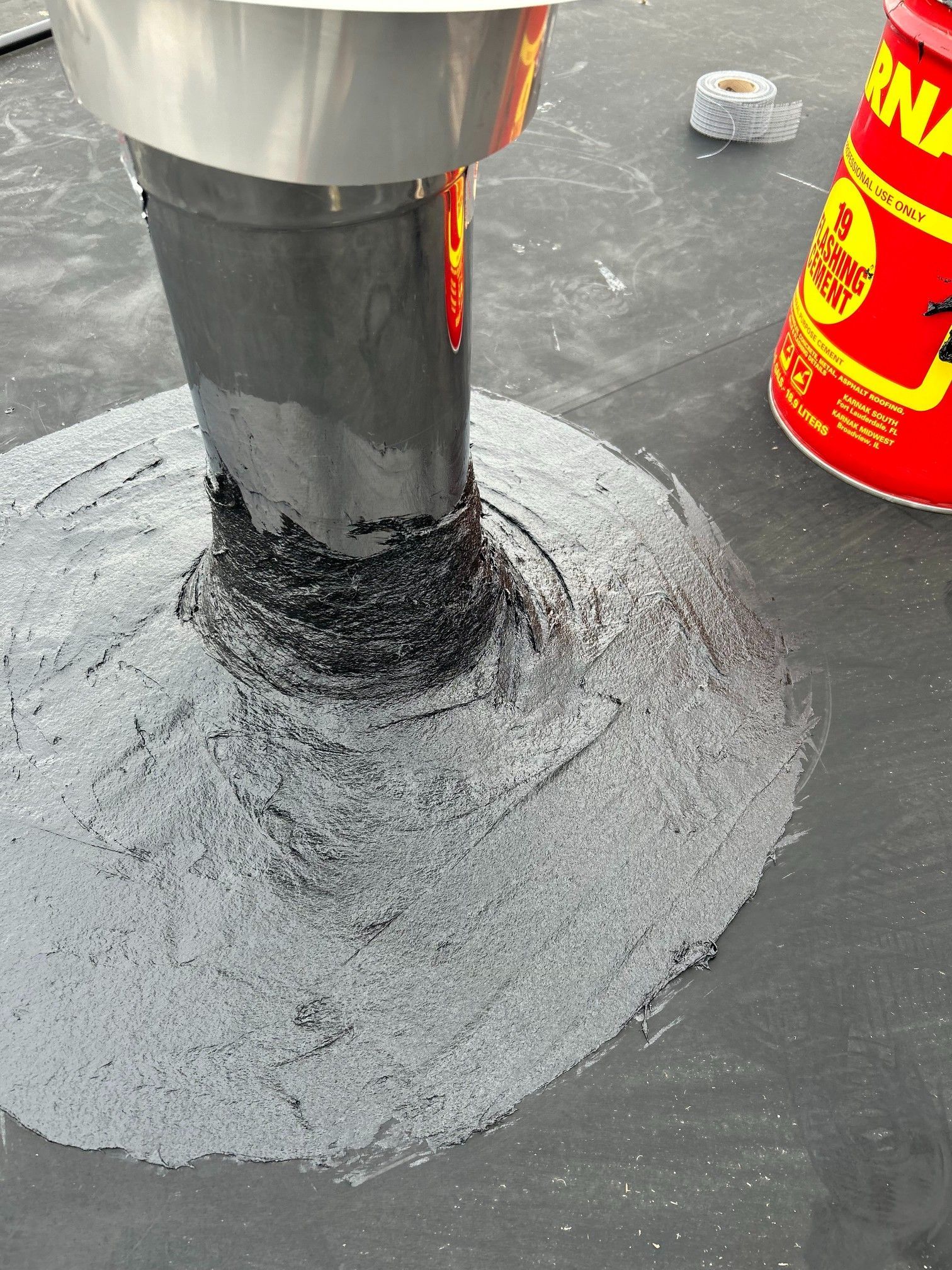 A black pipe sealed to a roof with a gray sealant and a red can of roofing sealant.