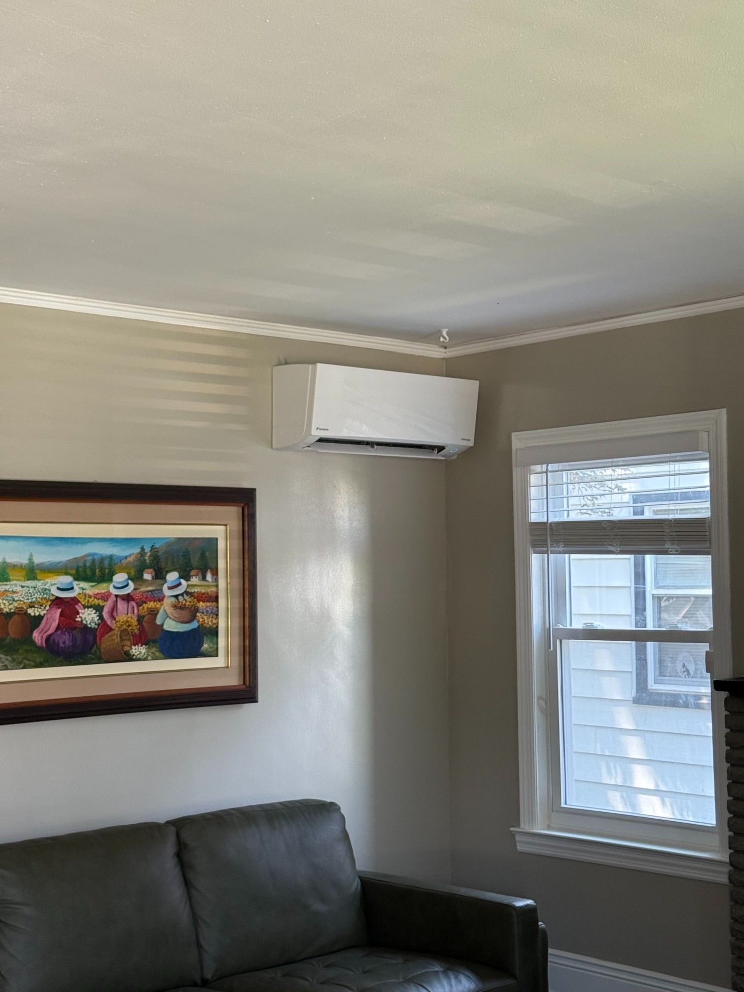 White wall-mounted air conditioner above a dark leather couch and a painting in a room with a window.
